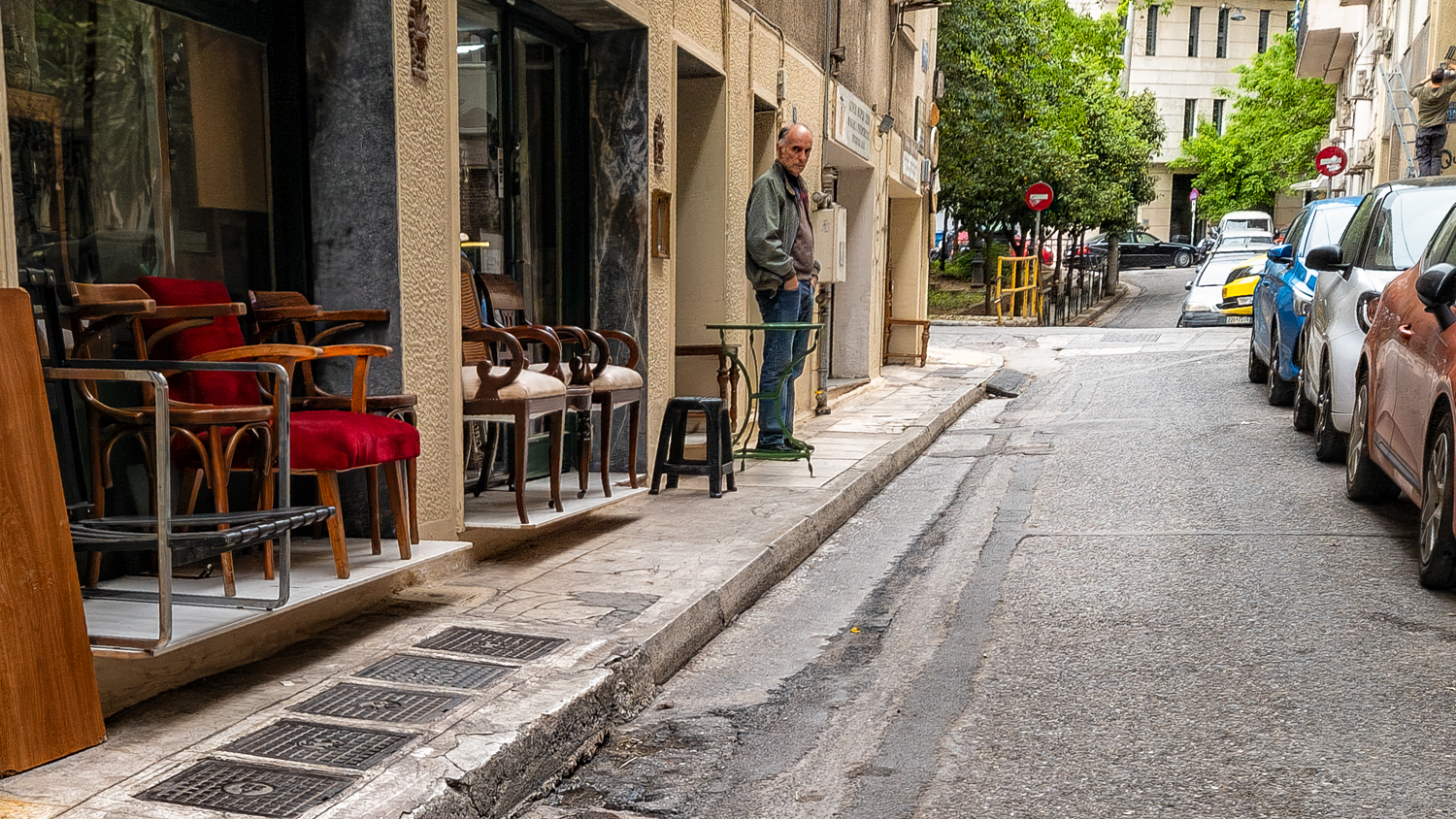 Athene: smalle straat met geparkeerde auto's, een rood stoeltje en een verkoper